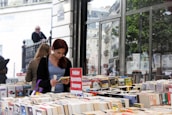 A person with red hair is absorbed in reading a book at an outdoor bookstall filled with a variety of books. A sign in French, indicating genres like crime and suspense, is displayed prominently. The background includes a large reflective glass window with trees and urban architecture visible, adding to the busy yet serene atmosphere of a street market.