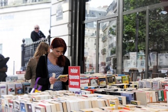 A person with red hair is absorbed in reading a book at an outdoor bookstall filled with a variety of books. A sign in French, indicating genres like crime and suspense, is displayed prominently. The background includes a large reflective glass window with trees and urban architecture visible, adding to the busy yet serene atmosphere of a street market.