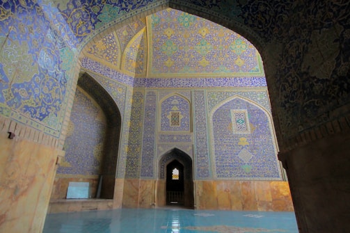Intricate Islamic architectural details featuring a richly decorated interior with ornate geometric patterns and calligraphy in blue and gold hues. High vaulted ceilings and archways create a majestic atmosphere.