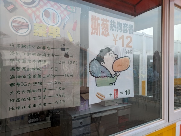 A window displays a menu with Chinese text and an advertisement poster showing a cartoon character holding a large sandwich. The text suggests prices for food items, including various Chinese dishes and a hot dog combo. The interior of a restaurant is visible through the glass, with tables, chairs, and some decorations.