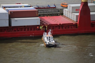 A large cargo ship is docked, loaded with multiple shipping containers stacked on its deck. A small pilot boat is maneuvering near the ship’s side in the water. Several people in safety gear stand on the ship's deck near the containers. In the background, there is a small orange vehicle parked on the dock.