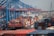 A bustling shipping terminal with numerous colorful shipping containers stacked on a large freight ship. The cranes and infrastructure towering above indicate an industrial environment, with the bridge in the background suggesting proximity to urban areas.