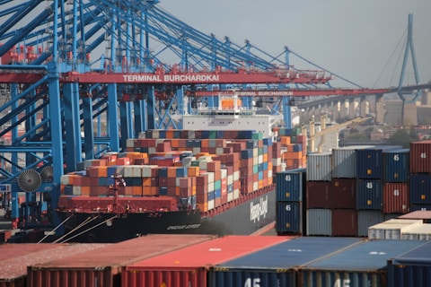 A bustling shipping terminal with numerous colorful shipping containers stacked on a large freight ship. The cranes and infrastructure towering above indicate an industrial environment, with the bridge in the background suggesting proximity to urban areas.