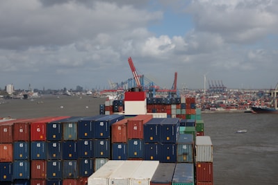 A large number of shipping containers are stacked on a cargo ship at a busy port. Several cranes are visible in the background, along with a view of the city skyline across the water. The sky is cloudy, and multiple ships and boats are navigating the harbor.