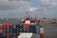 A large number of shipping containers are stacked on a cargo ship at a busy port. Several cranes are visible in the background, along with a view of the city skyline across the water. The sky is cloudy, and multiple ships and boats are navigating the harbor.