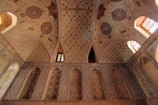 An intricately decorated ceiling and wall featuring geometric patterns and floral motifs. The design includes rich, earthy tones and detailed, symmetrical art. Arched windows allow warm light to illuminate the space, enhancing the ornate craftsmanship.