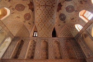 An intricately decorated ceiling and wall featuring geometric patterns and floral motifs. The design includes rich, earthy tones and detailed, symmetrical art. Arched windows allow warm light to illuminate the space, enhancing the ornate craftsmanship.
