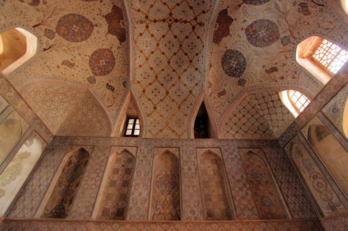 An intricately decorated ceiling and wall featuring geometric patterns and floral motifs. The design includes rich, earthy tones and detailed, symmetrical art. Arched windows allow warm light to illuminate the space, enhancing the ornate craftsmanship.