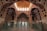 A richly decorated interior featuring intricate architectural details and ornate patterns on the walls and ceiling. The space is lined with glass partitions, allowing natural light to filter through large arched windows at the far end. The design features geometric and floral motifs with a warm, earthy color palette.