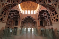 A richly decorated interior featuring intricate architectural details and ornate patterns on the walls and ceiling. The space is lined with glass partitions, allowing natural light to filter through large arched windows at the far end. The design features geometric and floral motifs with a warm, earthy color palette.