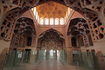 A richly decorated interior featuring intricate architectural details and ornate patterns on the walls and ceiling. The space is lined with glass partitions, allowing natural light to filter through large arched windows at the far end. The design features geometric and floral motifs with a warm, earthy color palette.