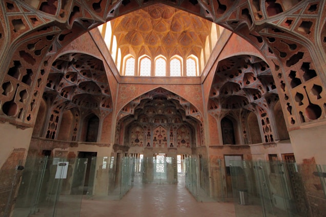 A richly decorated interior featuring intricate architectural details and ornate patterns on the walls and ceiling. The space is lined with glass partitions, allowing natural light to filter through large arched windows at the far end. The design features geometric and floral motifs with a warm, earthy color palette.
