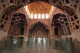 A richly decorated interior featuring intricate architectural details and ornate patterns on the walls and ceiling. The space is lined with glass partitions, allowing natural light to filter through large arched windows at the far end. The design features geometric and floral motifs with a warm, earthy color palette.