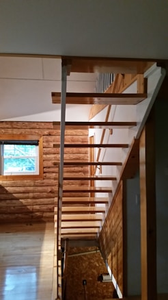 A wooden staircase with open risers leads up to a second floor, set against a backdrop of wooden walls and a window. The natural wood tones create a cabin-like atmosphere, with light streaming in from the window, casting shadows on the floor.