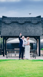 A couple is standing together in front of a rustic pavilion structure. The man is waving, dressed in a dark suit and light tie, while the woman wears a sparkling dress. A bird is flying above the pavilion in a cloudy sky. The foreground features a well-maintained lawn.