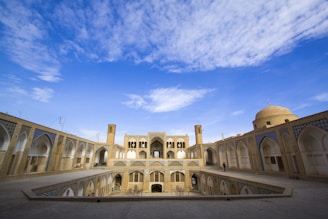 An architectural structure with arched doorways and intricate design patterns, set under a vast blue sky with scattered clouds. The building features a large courtyard surrounded by two stories of arcades. A domed structure is visible on the right, and tall windcatchers are present in the center, typical of traditional Persian architecture.