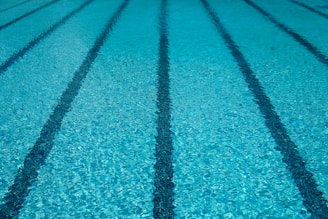 Clear blue water in a swimming pool with evenly spaced lane markers running across the length of the pool. The sunlight creates a shimmering effect on the water surface.