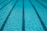 Clear blue water in a swimming pool with evenly spaced lane markers running across the length of the pool. The sunlight creates a shimmering effect on the water surface.