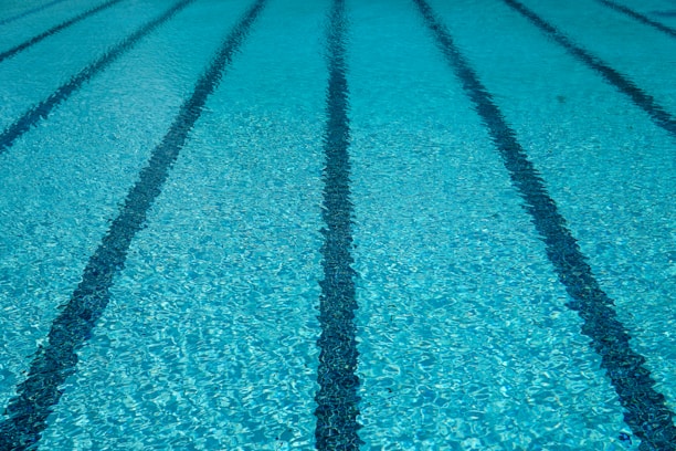 Clear blue water in a swimming pool with evenly spaced lane markers running across the length of the pool. The sunlight creates a shimmering effect on the water surface.