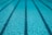 Clear blue water in a swimming pool with evenly spaced lane markers running across the length of the pool. The sunlight creates a shimmering effect on the water surface.