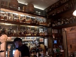 A dimly lit bar features an extensive collection of liquor bottles on wooden shelves, with warm lighting accentuating their presence. A bartender is seen working behind the counter, engaged with a device. Wooden barrels are placed on the upper shelf, contributing to a rustic ambiance.