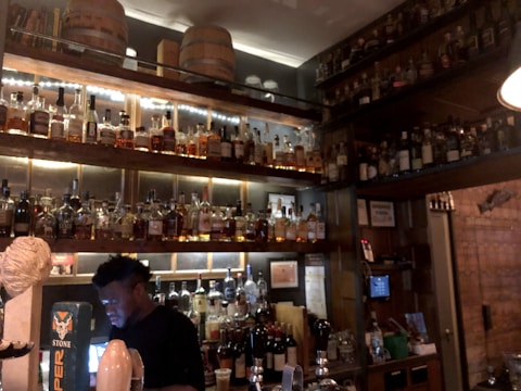A dimly lit bar features an extensive collection of liquor bottles on wooden shelves, with warm lighting accentuating their presence. A bartender is seen working behind the counter, engaged with a device. Wooden barrels are placed on the upper shelf, contributing to a rustic ambiance.