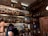 A dimly lit bar features an extensive collection of liquor bottles on wooden shelves, with warm lighting accentuating their presence. A bartender is seen working behind the counter, engaged with a device. Wooden barrels are placed on the upper shelf, contributing to a rustic ambiance.