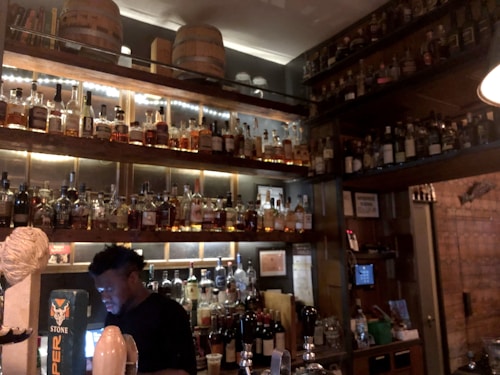 A dimly lit bar features an extensive collection of liquor bottles on wooden shelves, with warm lighting accentuating their presence. A bartender is seen working behind the counter, engaged with a device. Wooden barrels are placed on the upper shelf, contributing to a rustic ambiance.
