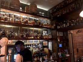 A dimly lit bar features an extensive collection of liquor bottles on wooden shelves, with warm lighting accentuating their presence. A bartender is seen working behind the counter, engaged with a device. Wooden barrels are placed on the upper shelf, contributing to a rustic ambiance.