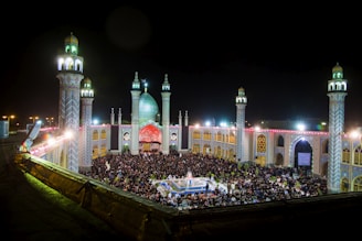 A large gathering of people is seen at night in an ornate courtyard, surrounded by beautifully lit minarets and domes. The architecture is intricate, with colorful patterns and lights illuminating the scene. A central fountain is visible among the crowd.