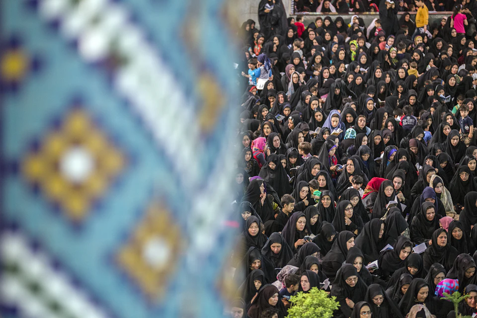 The image depicts a large crowd of people gathered together, all dressed in black clothing and facing towards the right side of the frame. The perspective is from above, providing an aerial view of the scene. In the background, there are buildings visible, suggesting that this gathering might be taking place in an urban setting or near a public space.