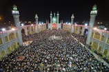 A large crowd of people gathered in an open courtyard surrounded by a mosque with four lit minarets at night. The architecture is ornate, featuring detailed tile work and archways, illuminated by bright lighting. The scene is vibrant and busy with people standing and seated closely together.
