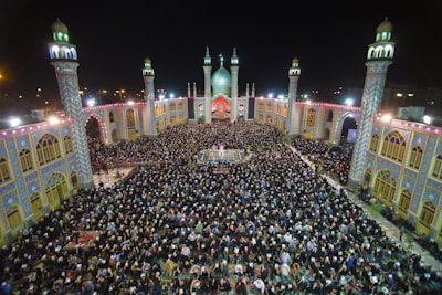 A large crowd of people gathered in an open courtyard surrounded by a mosque with four lit minarets at night. The architecture is ornate, featuring detailed tile work and archways, illuminated by bright lighting. The scene is vibrant and busy with people standing and seated closely together.