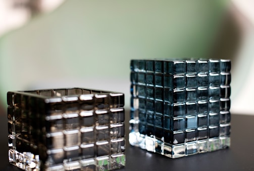 Two glass cubes with a grid pattern sit on a dark surface. The cubes appear transparent with a subtle reflection of light, one slightly tinted darker than the other. The background is blurred with soft green and gray tones.
