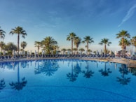 Private swimming pool reflecting the clear blue sky, framed by sandy tones and greenery.