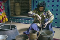 An elderly person in traditional clothing is seated on the floor, stirring the contents of a metal pot with a large spoon. The background features decorative blue tiles and a clay jug rests on a brick shelf.