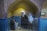 Three mannequins are dressed in traditional clothing within a small alcove. The alcove features brickwork arches and blue-tiled walls. There is a framed picture on one side of the wall, and the lighting casts a warm glow, highlighting the textures of the bricks.