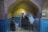 Three mannequins are dressed in traditional clothing within a small alcove. The alcove features brickwork arches and blue-tiled walls. There is a framed picture on one side of the wall, and the lighting casts a warm glow, highlighting the textures of the bricks.