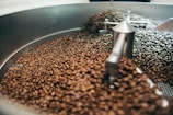 A large quantity of coffee beans inside an industrial roasting machine. The beans are in motion, possibly being stirred or cooled, and are enclosed by a metallic circular container with various metal parts visible.