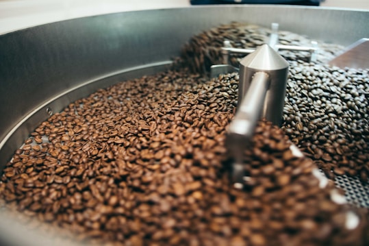 A large quantity of coffee beans inside an industrial roasting machine. The beans are in motion, possibly being stirred or cooled, and are enclosed by a metallic circular container with various metal parts visible.