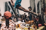 A vibrant street scene with a market setup featuring various jars, bottles, and baskets of food items displayed on a table. In the background, a mural depicts a crowd of people and a dancer in mid-air with an arched back. A person wearing a patterned outfit and a red headscarf is seen from behind, possibly attending to the stall.