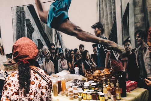 A vibrant street scene with a market setup featuring various jars, bottles, and baskets of food items displayed on a table. In the background, a mural depicts a crowd of people and a dancer in mid-air with an arched back. A person wearing a patterned outfit and a red headscarf is seen from behind, possibly attending to the stall.