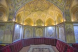 An intricately decorated room with an arched ceiling adorned with detailed motifs and patterns. The walls are embellished with elaborate floral designs and the room features red cushioned seating along the perimeter. A patterned carpet is placed at the center of the room.