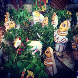 A collection of colorful clay idols, predominantly featuring human figures with painted clothing and expressions. The idols are surrounded by fresh green leaves and small flowers. A white painted cow figurine with decorative patterns is prominently visible among the idols.