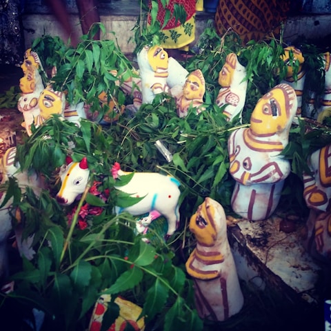 A collection of colorful clay idols, predominantly featuring human figures with painted clothing and expressions. The idols are surrounded by fresh green leaves and small flowers. A white painted cow figurine with decorative patterns is prominently visible among the idols.