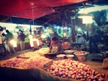 A bustling marketplace scene featuring a variety of onions displayed on tables and in baskets. Vendors and customers are interacting under bright artificial lights. The scene is lively with people standing and moving around, indicating a busy market atmosphere.