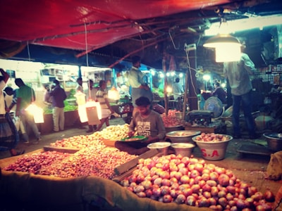 A bustling marketplace scene featuring a variety of onions displayed on tables and in baskets. Vendors and customers are interacting under bright artificial lights. The scene is lively with people standing and moving around, indicating a busy market atmosphere.