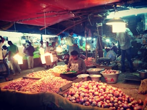A bustling marketplace scene featuring a variety of onions displayed on tables and in baskets. Vendors and customers are interacting under bright artificial lights. The scene is lively with people standing and moving around, indicating a busy market atmosphere.