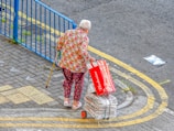 An elderly person with white hair is walking with the aid of a yellow cane. They are dressed in colorful clothing featuring a floral shirt and patterned pants. They are pulling a small cart filled with bundled newspapers. The person is also holding an empty Budweiser beer box. The scene is set on a paved area next to a blue metal fence with a yellow road marking curving alongside.