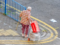 An elderly person with white hair is walking with the aid of a yellow cane. They are dressed in colorful clothing featuring a floral shirt and patterned pants. They are pulling a small cart filled with bundled newspapers. The person is also holding an empty Budweiser beer box. The scene is set on a paved area next to a blue metal fence with a yellow road marking curving alongside.